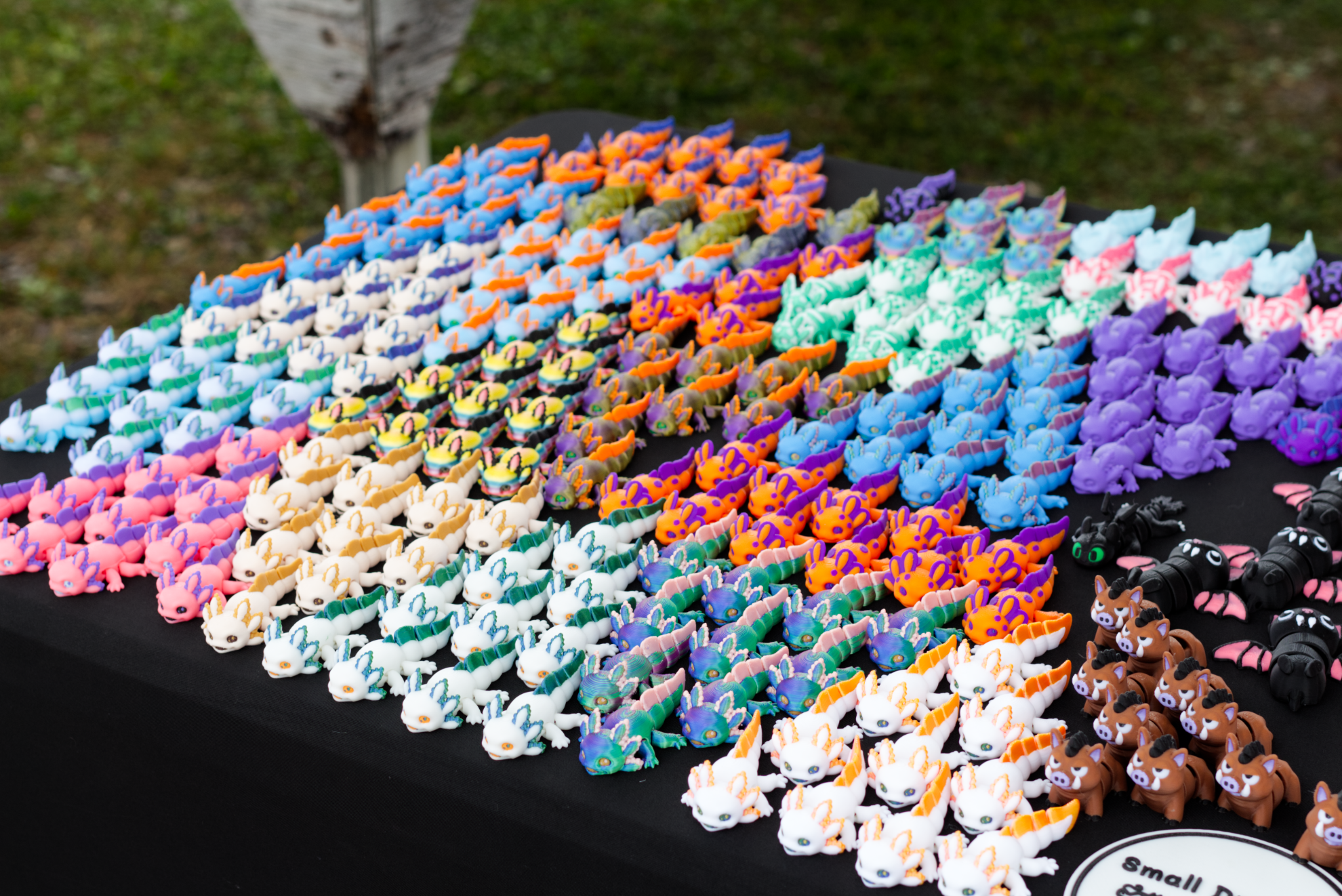 A table with many small 3D printed axolotls in different colors as well as a small black dragon, wild boars, and bats